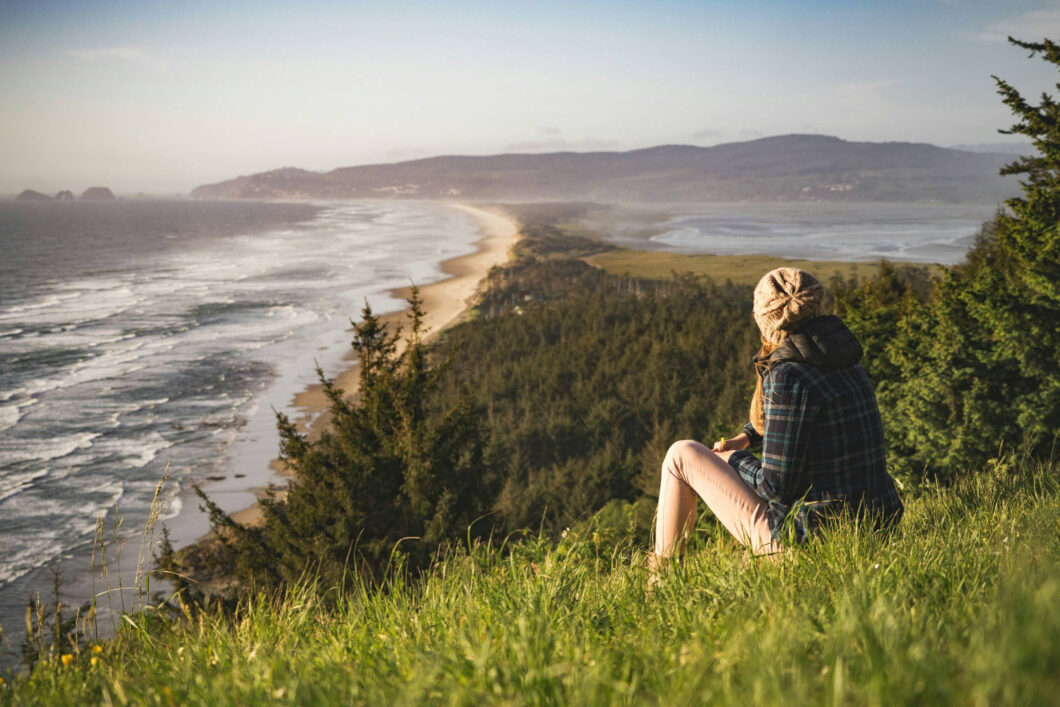 A person wearing a beanie and plaid shirt sits on a grassy hill overlooking a scenic coastline, reflecting on awareness in binge eating recovery amid waves, forest, and distant mountains under a clear sky.