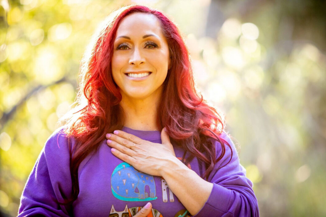 Kristina Dobyns smiling warmly outdoors, wearing a purple sweatshirt and holding one hand to her chest—a moment of peace on her food addiction recovery journey. Sunlight filters through the trees in the blurred background.