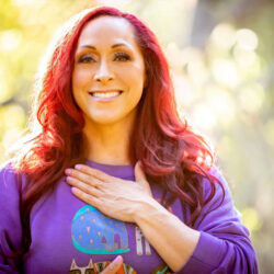 Kristina Dobyns smiling warmly outdoors, wearing a purple sweatshirt and holding one hand to her chest—a moment of peace on her food addiction recovery journey. Sunlight filters through the trees in the blurred background.