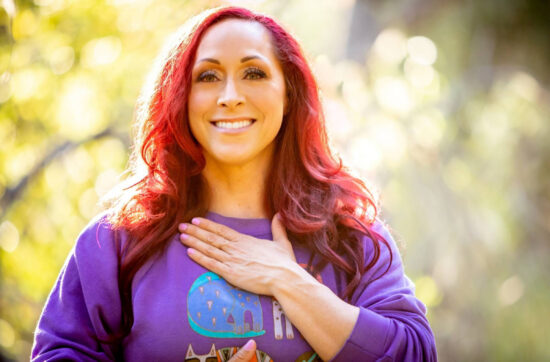 Kristina Dobyns smiling warmly outdoors, wearing a purple sweatshirt and holding one hand to her chest—a moment of peace on her food addiction recovery journey. Sunlight filters through the trees in the blurred background.
