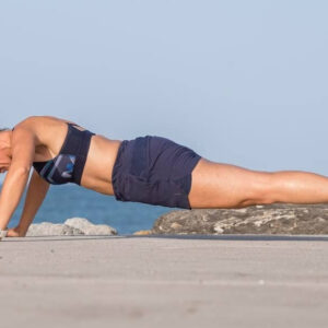 A woman in athletic wear demonstrates proprioceptive grounding techniques with a plank exercise outdoors on a concrete surface, set against the ocean and blue sky in the background.